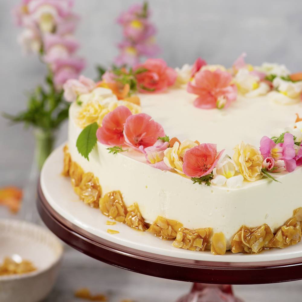 Runder Kuchen mit heller Creme, dekoriert mit bunten Blüten und Karamellstücken auf einem glänzenden Teller. Im Hintergrund Blumen.