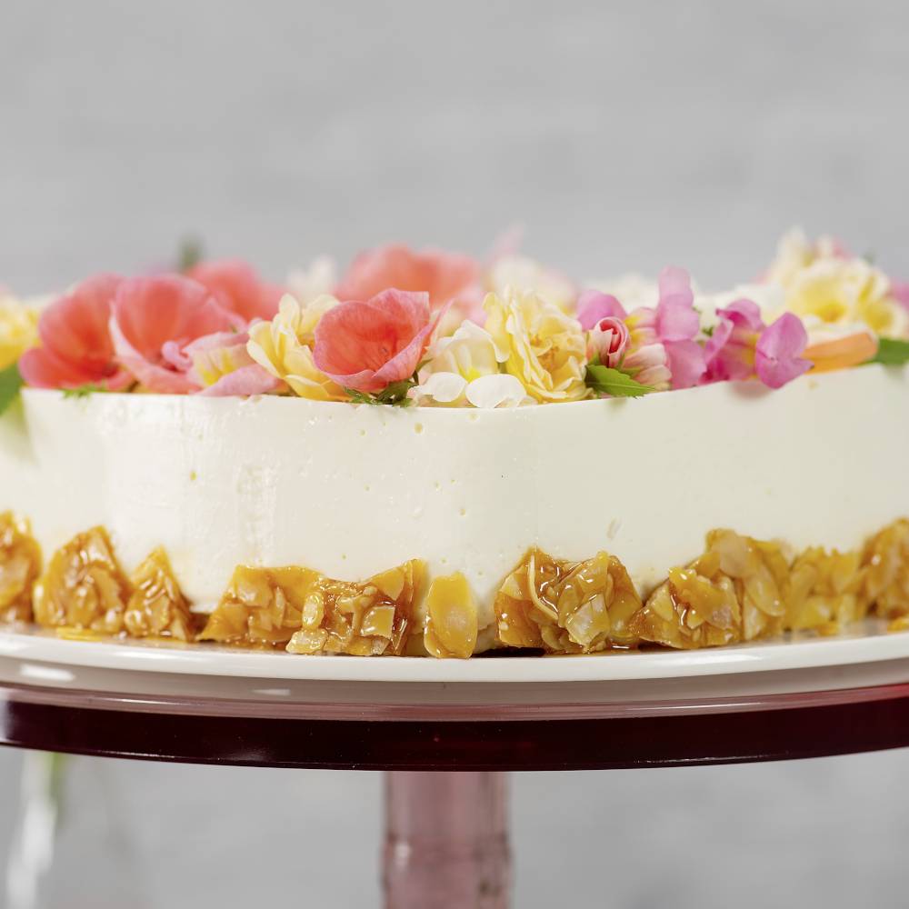 Ein runder Kuchen mit cremeweißer Glasur, dekoriert mit roten und gelben Blumen oben und kandierten Nüssen an der Seite.