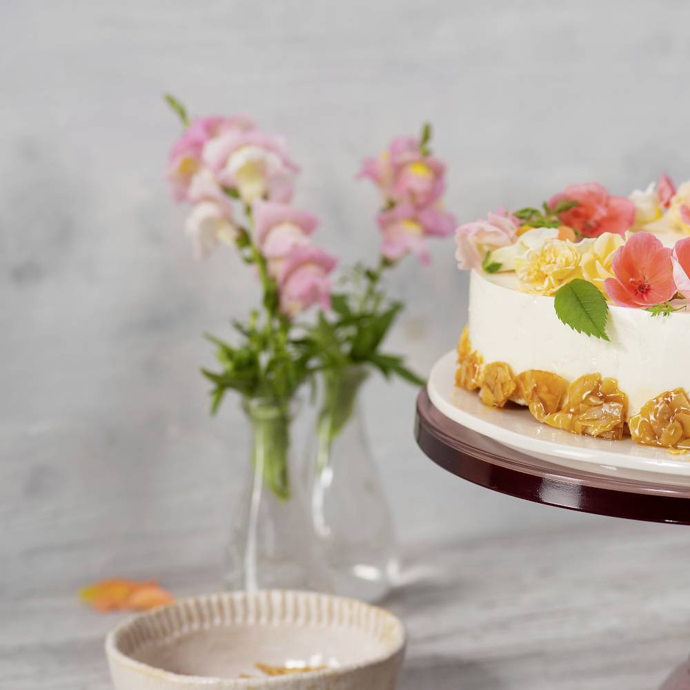 Ein Kuchen auf einem Tortenteller dekoriert mit rosa und orangefarbenen Blumen. Dahinter steht eine Vase mit ähnlichen Blumen.
