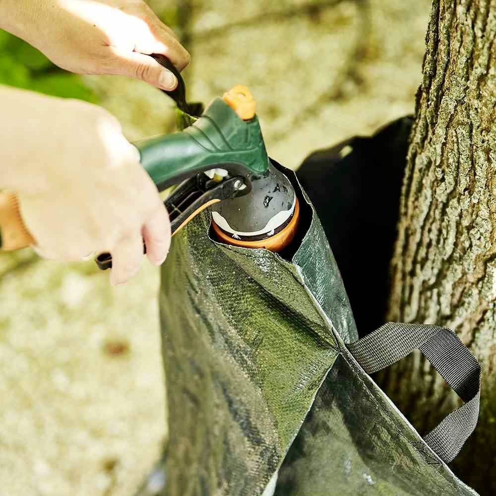 Person gießt einen Baum mit einer Gießkanne. Eine grüne Schutzhülle umgibt den Baumstamm, um Wasser zu halten.