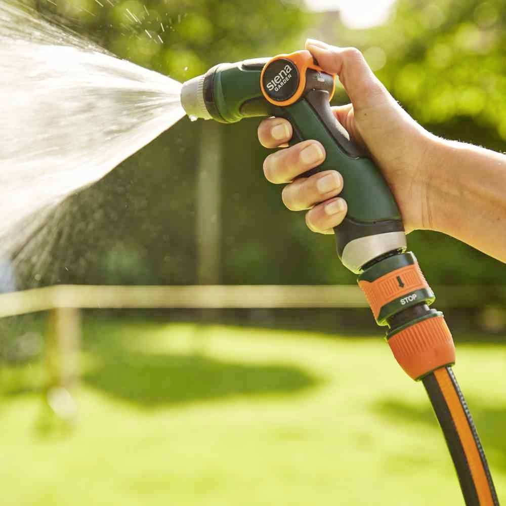 Eine Hand hält einen Gartenschlauch, der Wasser sprüht. Der Hintergrund zeigt einen unscharfen, grünen Garten.