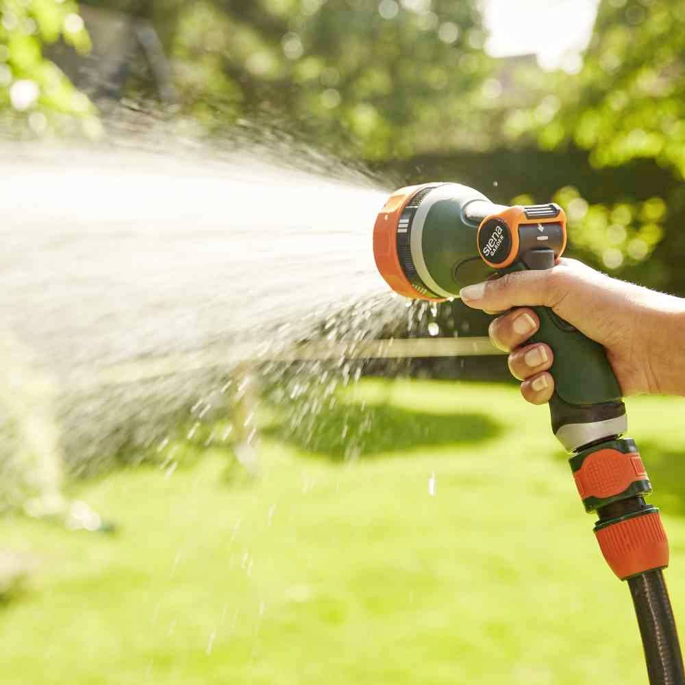 Eine Hand hält eine Gartenspritzpistole, die Wasser in einem gebogenen Strahl auf eine sonnige grüne Rasenfläche sprüht.
