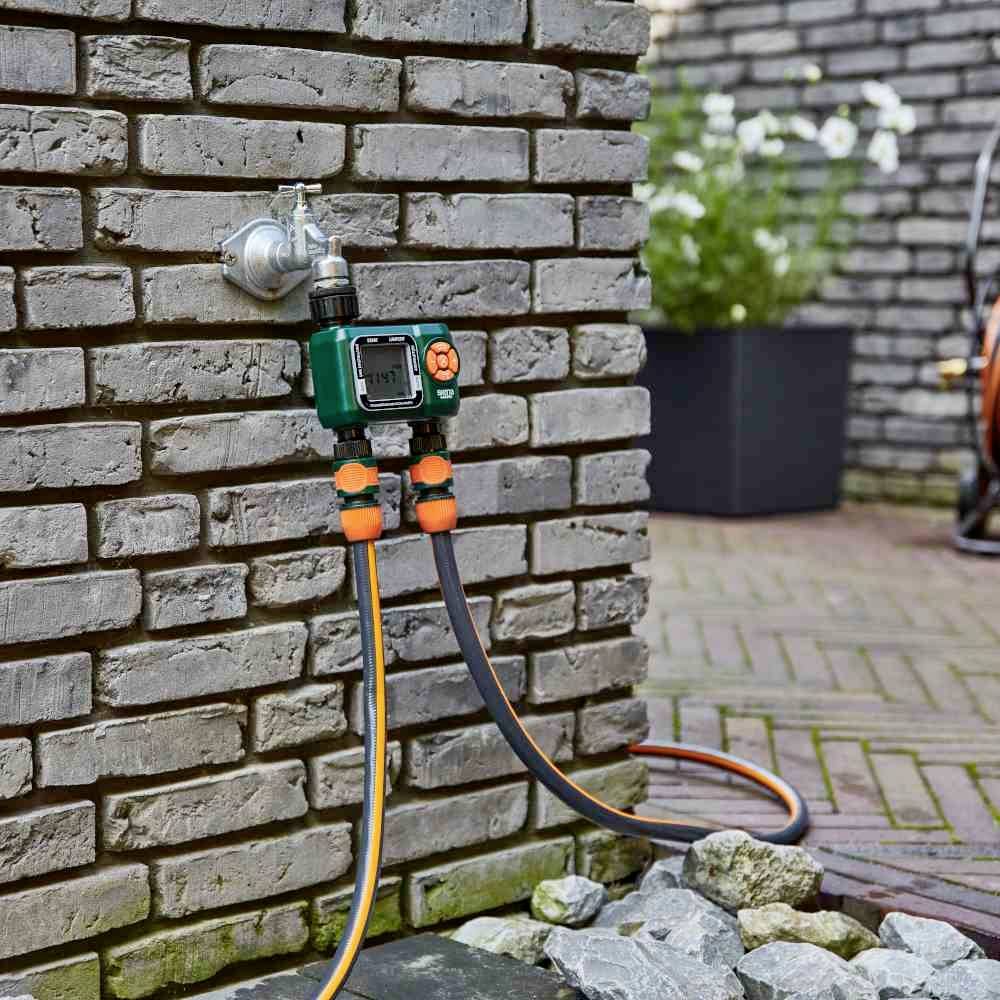 Wasserhahn an einer Ziegelmauer mit angeschlossenem Bewässerungssystem, zwei Schläuche. Hintergrund: Terrasse mit Pflanzen.
