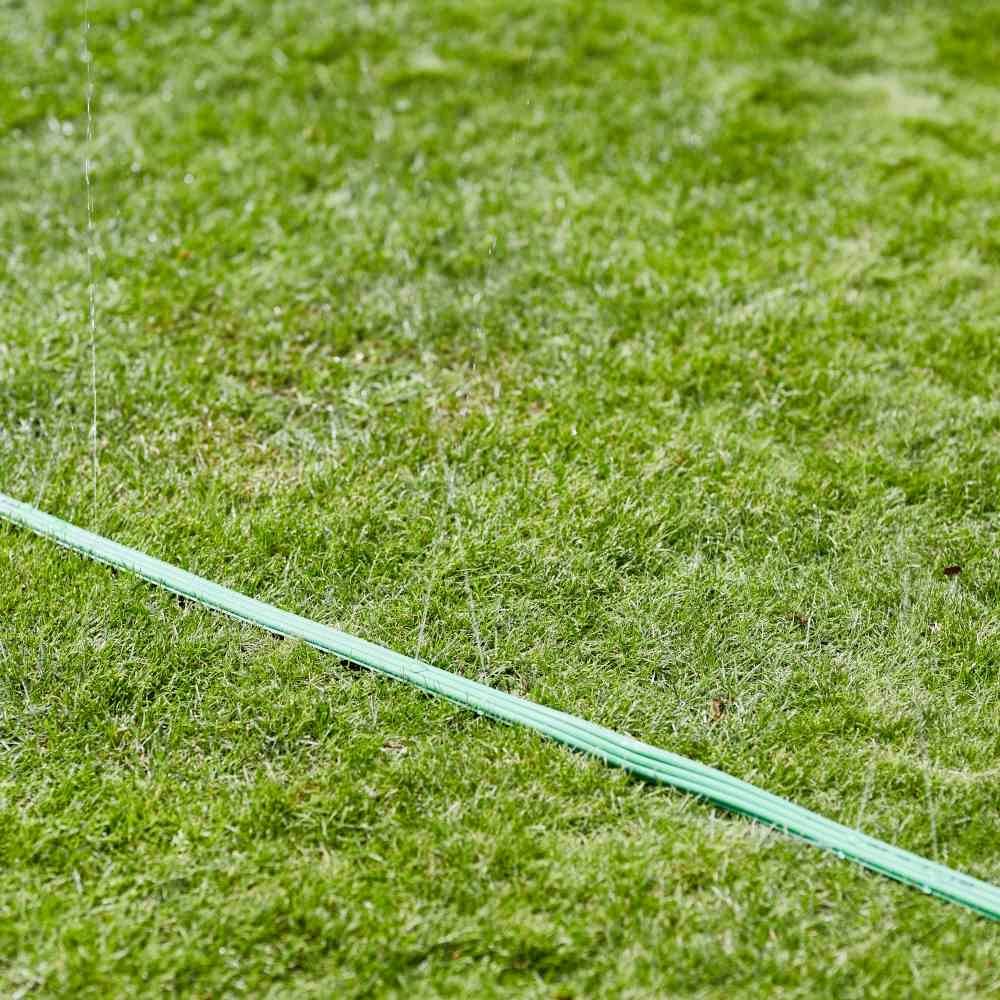 Ein grüner Gartenschlauch liegt auf einer Wiese und sprüht einen feinen Wassernebel, der das Gras befeuchtet.
