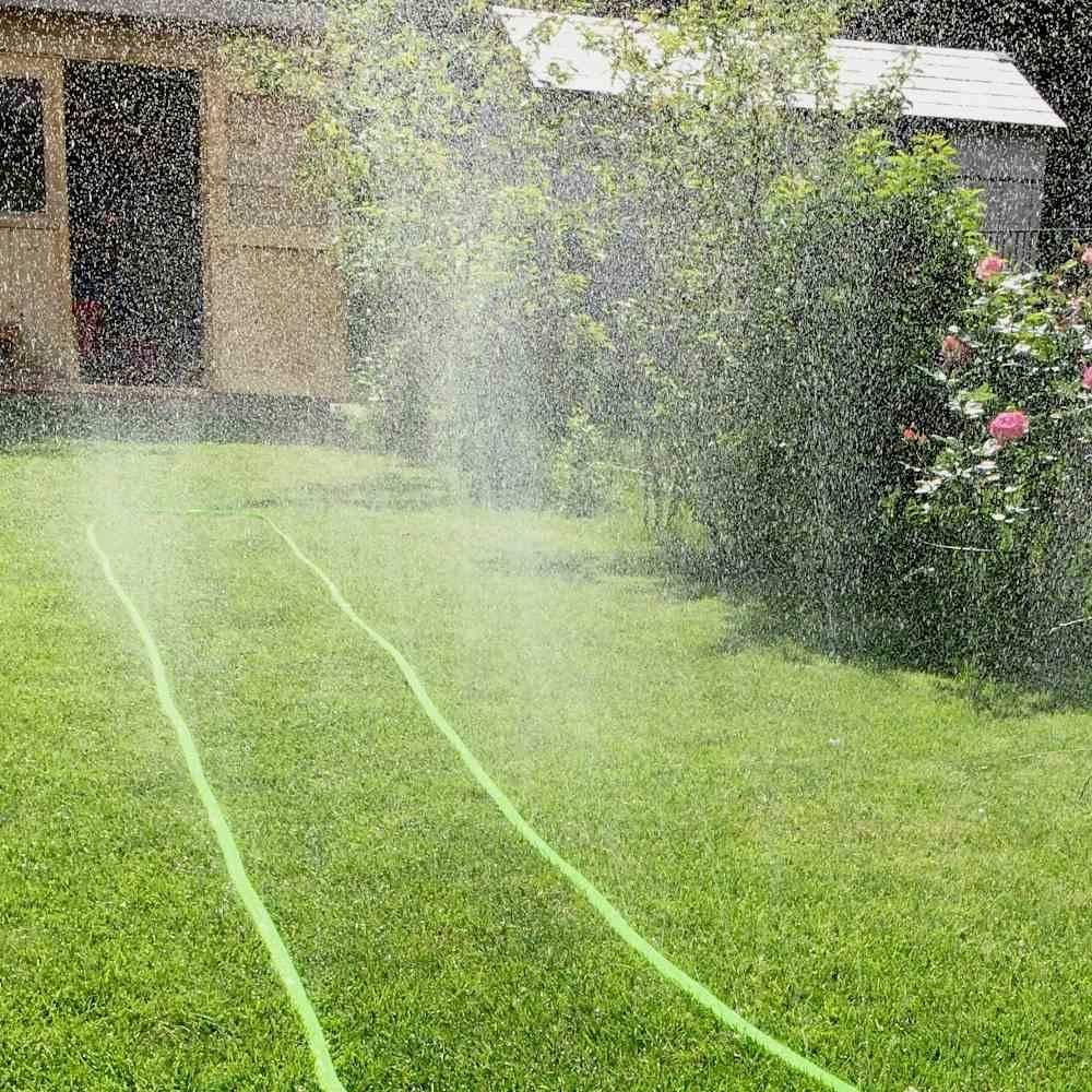 Rasen mit Rasensprenger, der fein verteiltes Wasser sprüht. Im Hintergrund ein kleiner Schuppen und blühende Sträucher.