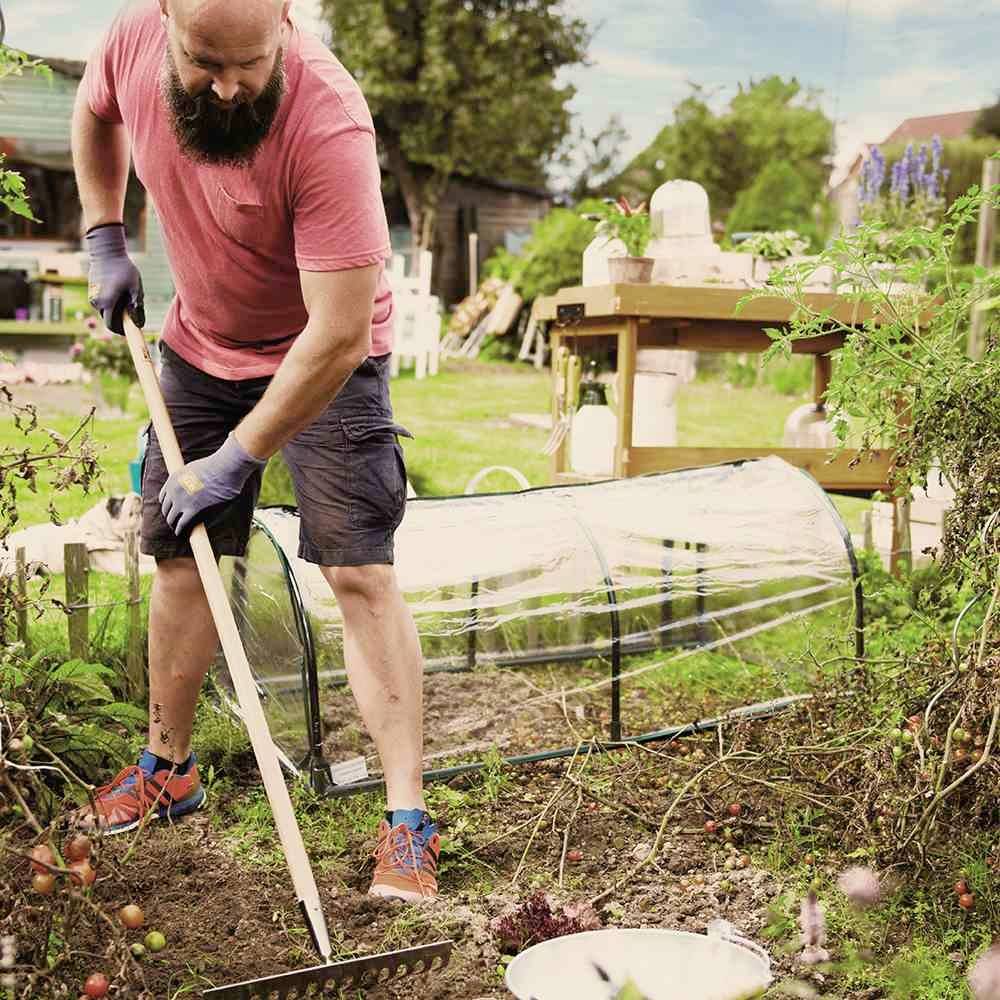 Eine Person mit Bart arbeitet im Garten, trägt rote Kleidung und bearbeitet den Boden mit einer Harke. Im Hintergrund sind Pflanzen und Gartenmöbel zu sehen.