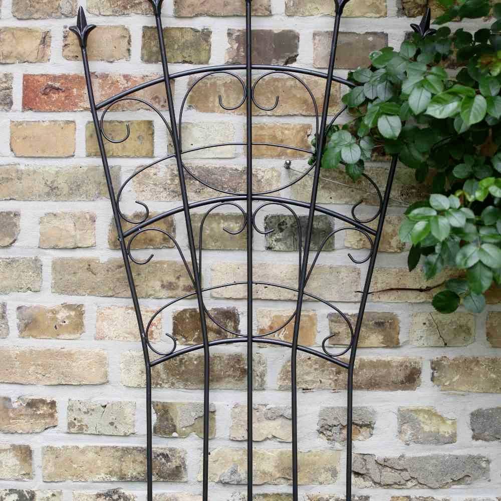 Ein schmiedeeiserner Pflanzenhalter steht vor einer Backsteinmauer, umgeben von grünen Blättern auf der rechten Seite.
