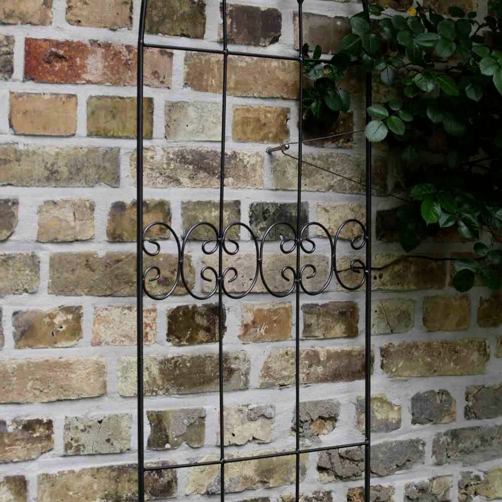 Ein schmiedeeiserner Gitterrost lehnt an einer Ziegelwand. Ein Rosenzweig windet sich durch das Gitterwerk.