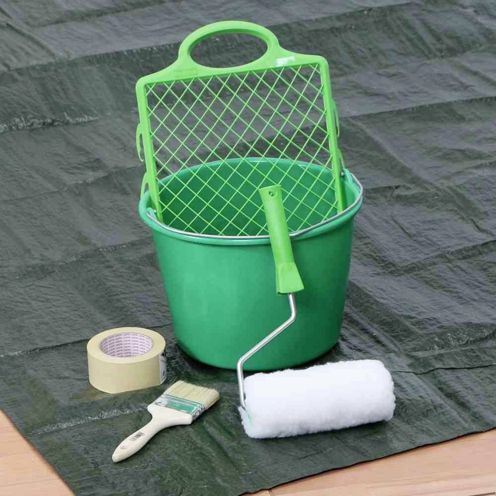 Ein grüner Eimer mit Farbgitter, ein Farbroller, ein Pinsel und ein Klebeband liegen auf einer Abdeckplane. Malzubehör.