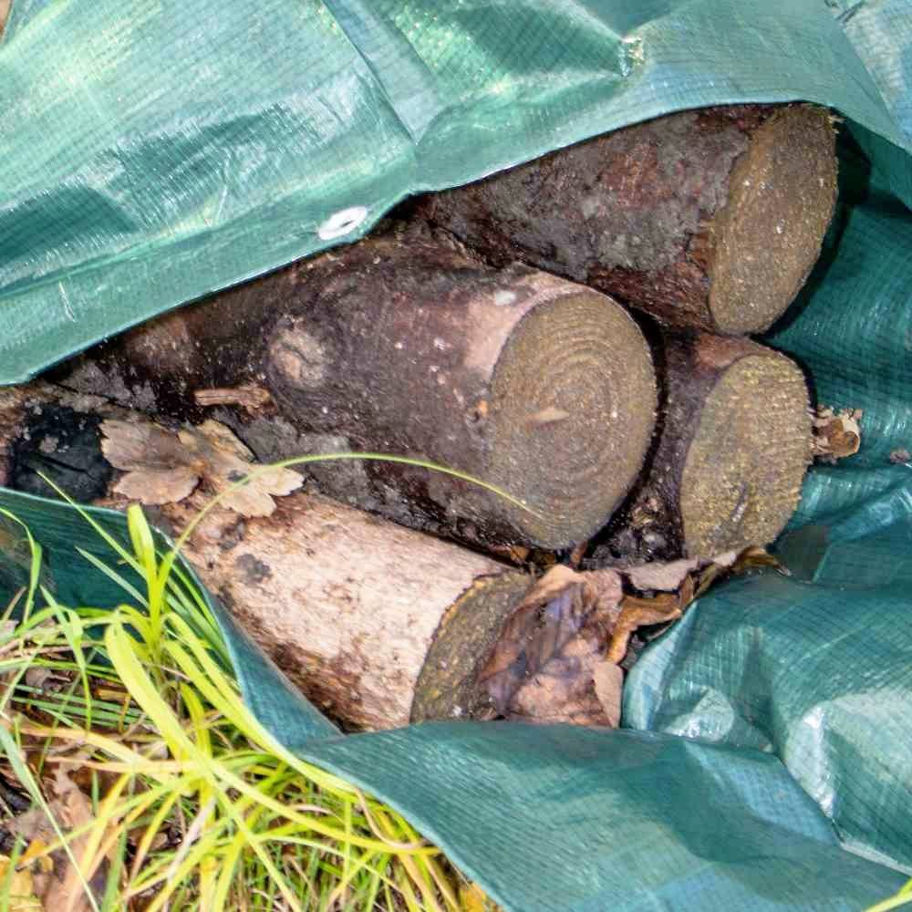 Holzscheite unter grüner Plane, teilweise von Gras bedeckt. Sie sind zum Trocknen gestapelt, bereit für den Wintergebrauch.