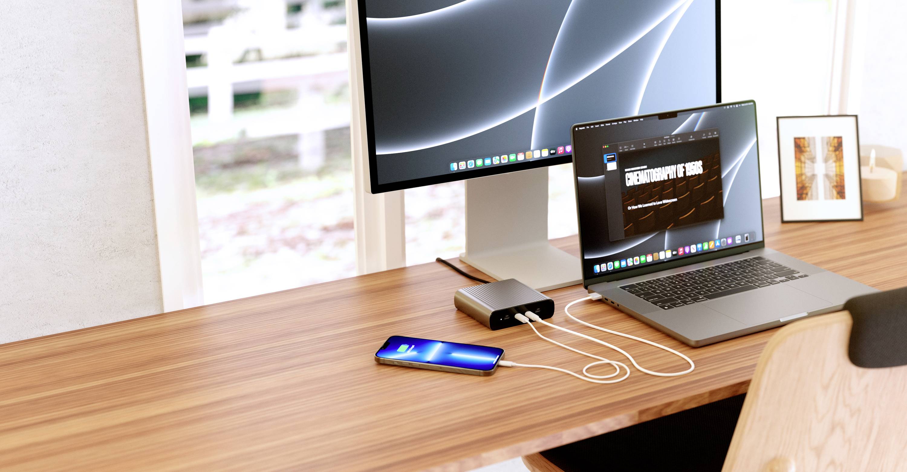 Arbeitsplatz mit Laptop und Smartphone, beide verbunden mit einem Ladegerät. Ein Monitor zeigt abstrakte Linien auf schwarzem Hintergrund.