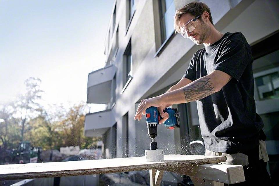 Ein Mann bohrt mit einer elektrischen Bohrmaschine ein Loch in ein Holzbrett auf einer Baustelle, trägt Sicherheitsbrille.