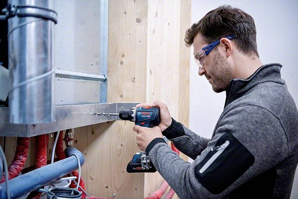 Ein Mann in Schutzbrille benutzt eine elektrische Bohrmaschine, um ein Loch in ein Metallstück zu bohren, das an einer Holzwand befestigt ist.