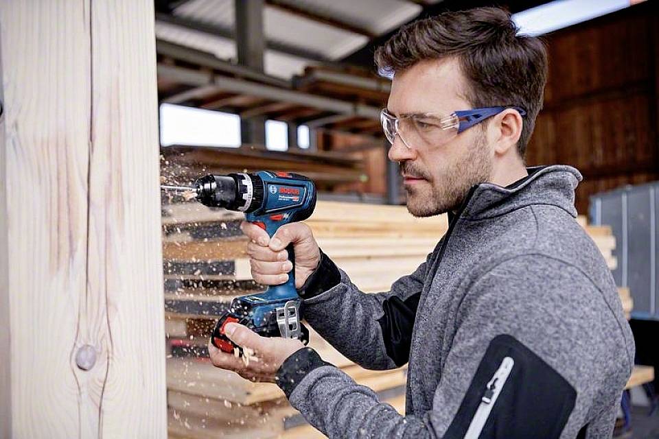 Ein Mann in Schutzbrille bohrt mit einer blauen Bohrmaschine in Holz. Er steht in einer Werkstatt mit mehreren Holzstapeln.