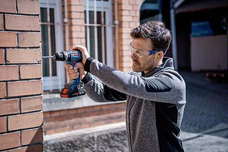 Ein Mann in Schutzbrille bohrt mit einem Akkubohrschrauber ein Loch in eine Ziegelwand. Im Hintergrund sind Fenster und Gebäude.