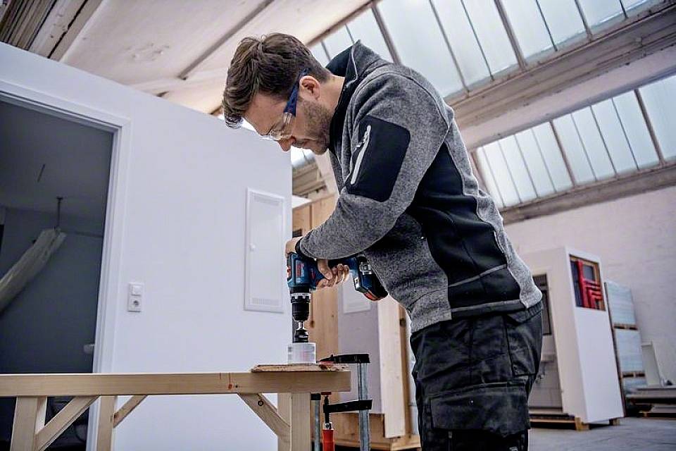 Ein Mann mit Schutzbrille benutzt eine elektrische Bohrmaschine, um in einem hellen Raum mit Dachfenstern Holz zu bearbeiten.