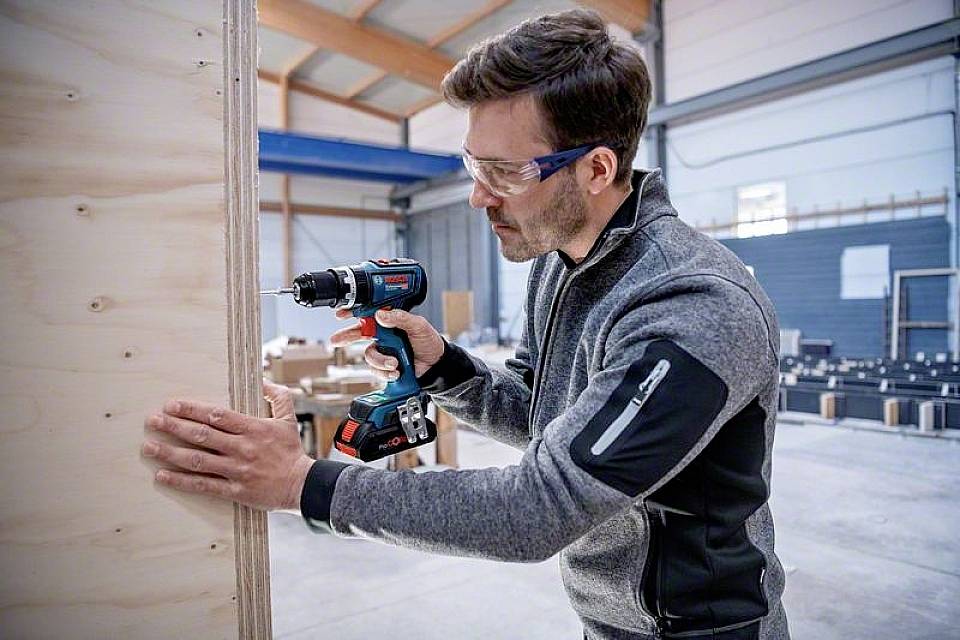 Ein Mann in Schutzbrille bohrt mit einer elektrischen Bohrmaschine in eine Holzplatte in einer Werkstatt oder Lagerhalle.