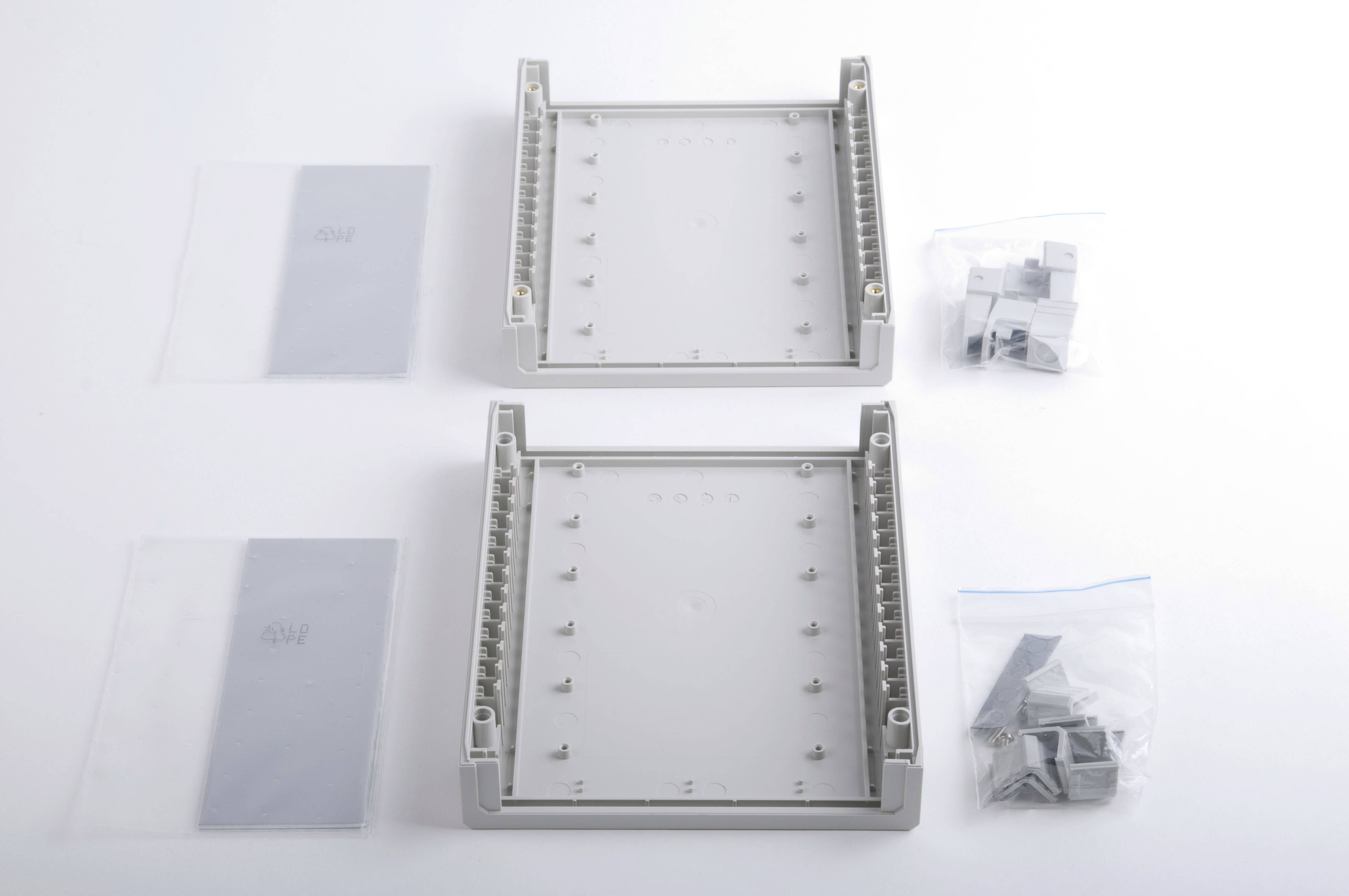 Zwei graue Kunststoffgehäuse liegen nebeneinander, umgeben von Verpackungen mit Schrauben und Montagezubehör auf weißem Hintergrund.