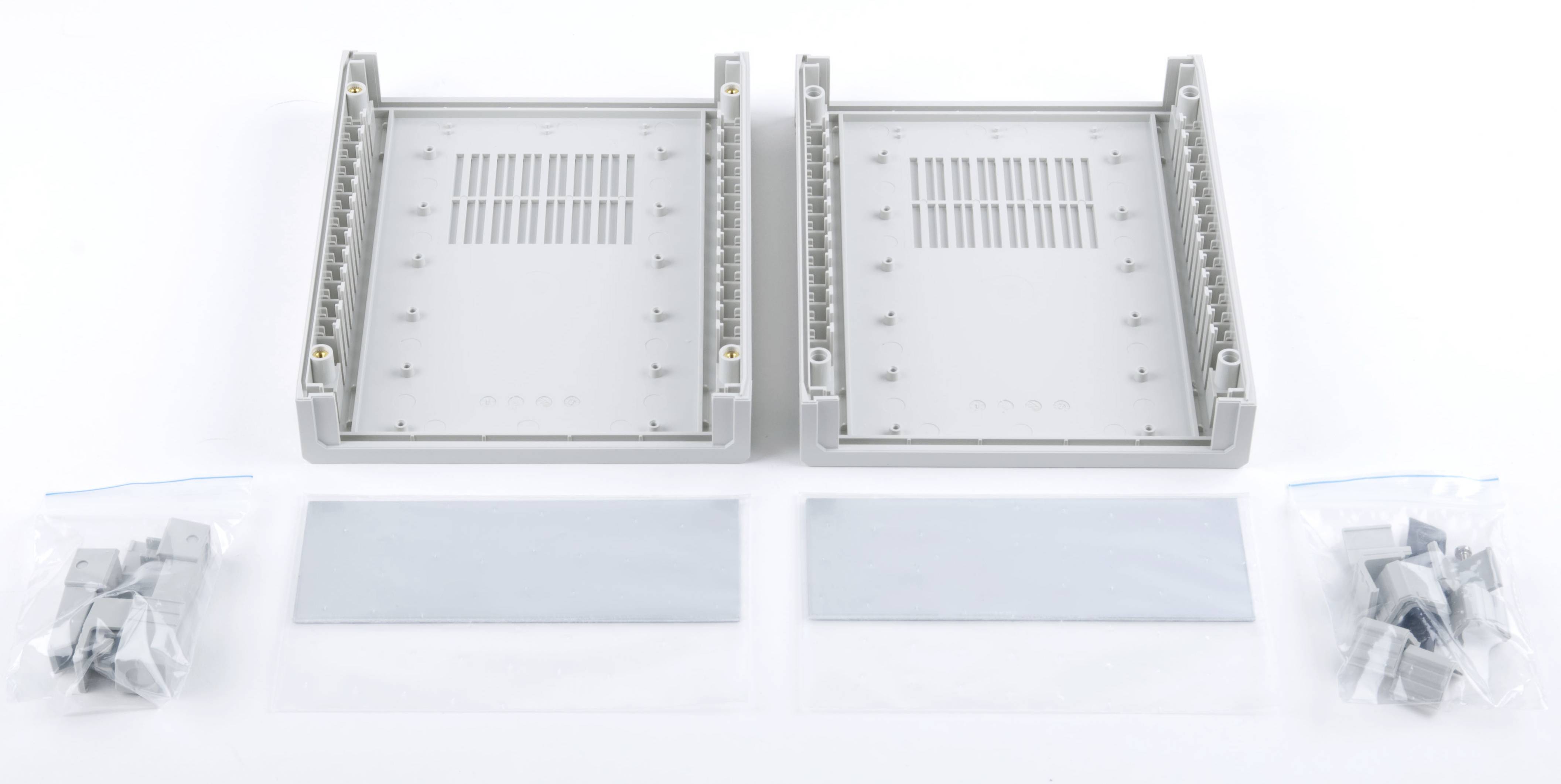 Zwei graue Projektboxen mit offenen Oberseiten, umgeben von kleinem Zubehör, darunter Steckverbinder und Kabelabdeckungen, auf weißem Hintergrund.