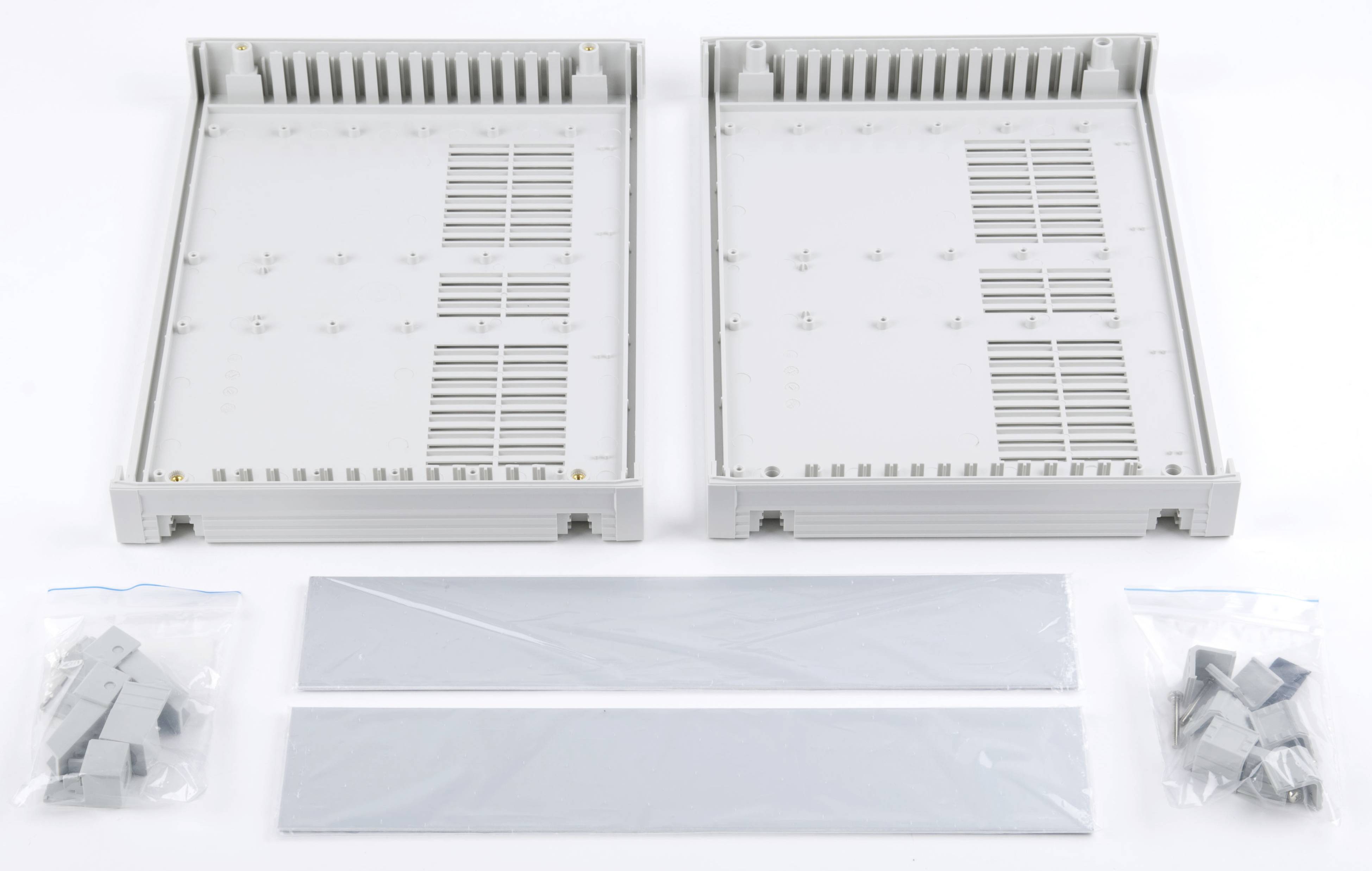 Zwei graue, rechteckige Kunststoffgehäuse mit Lüftungsschlitzen liegen offen. Daneben liegen zwei schmale Abdeckungen und kleine Klemmen in Plastiktüten.