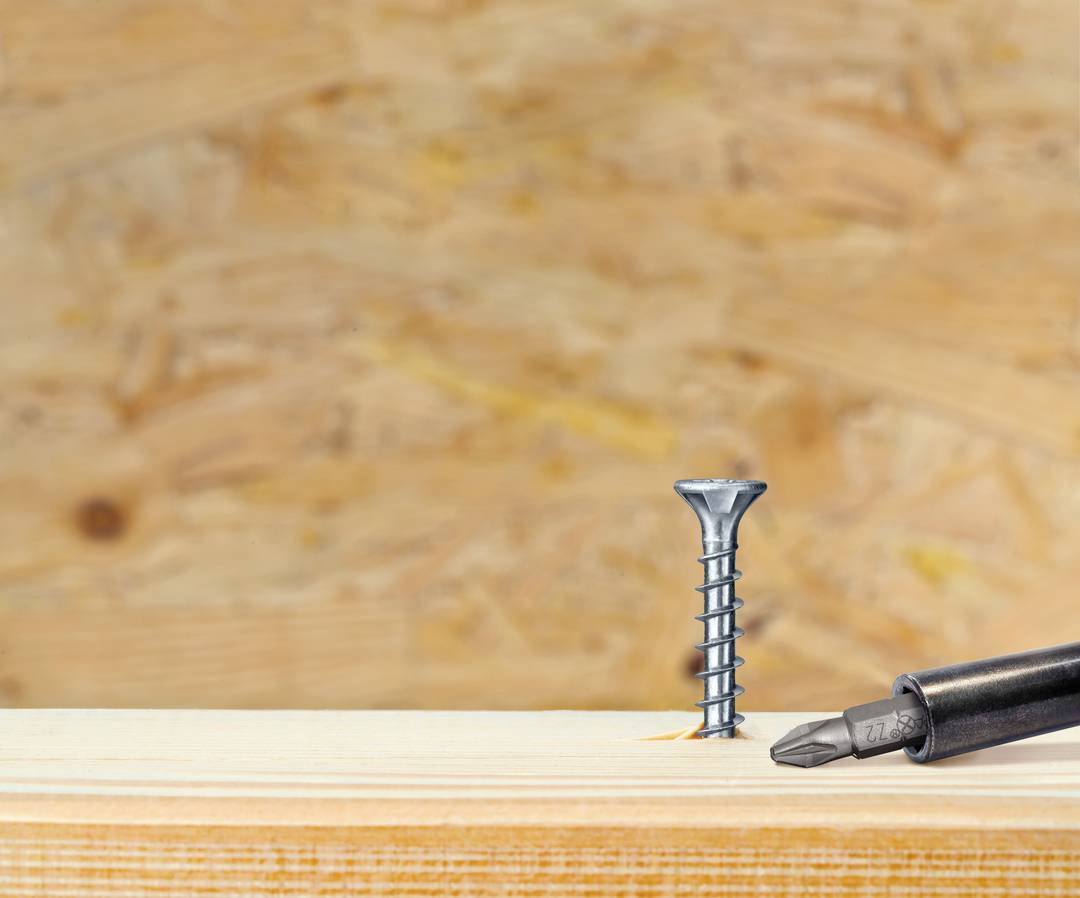 Ein Schraubenzieher liegt neben einer senkrecht auf einer Holzoberfläche stehenden Schraube, vor einem Hintergrund aus Sperrholz.