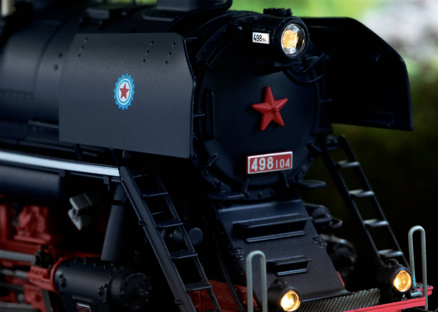 Schwarze Dampflokomotive mit rotem Sternemblem und der Nummer '498.104' vorne. Beleuchtete Scheinwerfer, umgeben von Bäumen.
