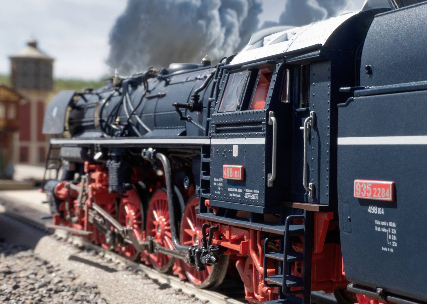 Eine Dampflokomotive fährt auf Schienen, mit weißem Rauch aus dem Schornstein. Im Hintergrund ist ein Gebäude zu sehen.
