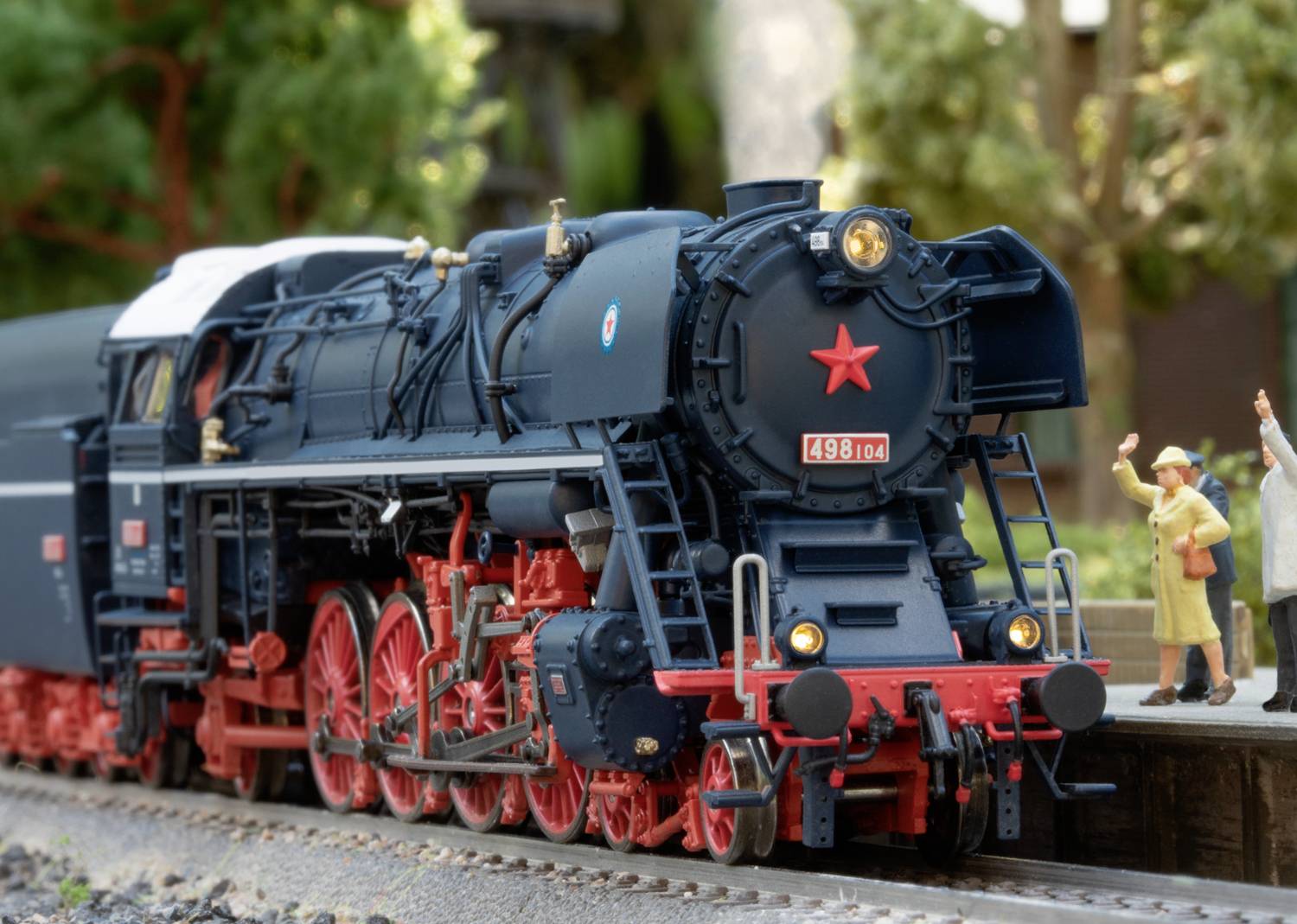 Eine schwarze Dampflokomotive mit roten Rädern fährt auf einem Gleis. Neben der Lokomotive stehen Figuren, die freundlich winken.