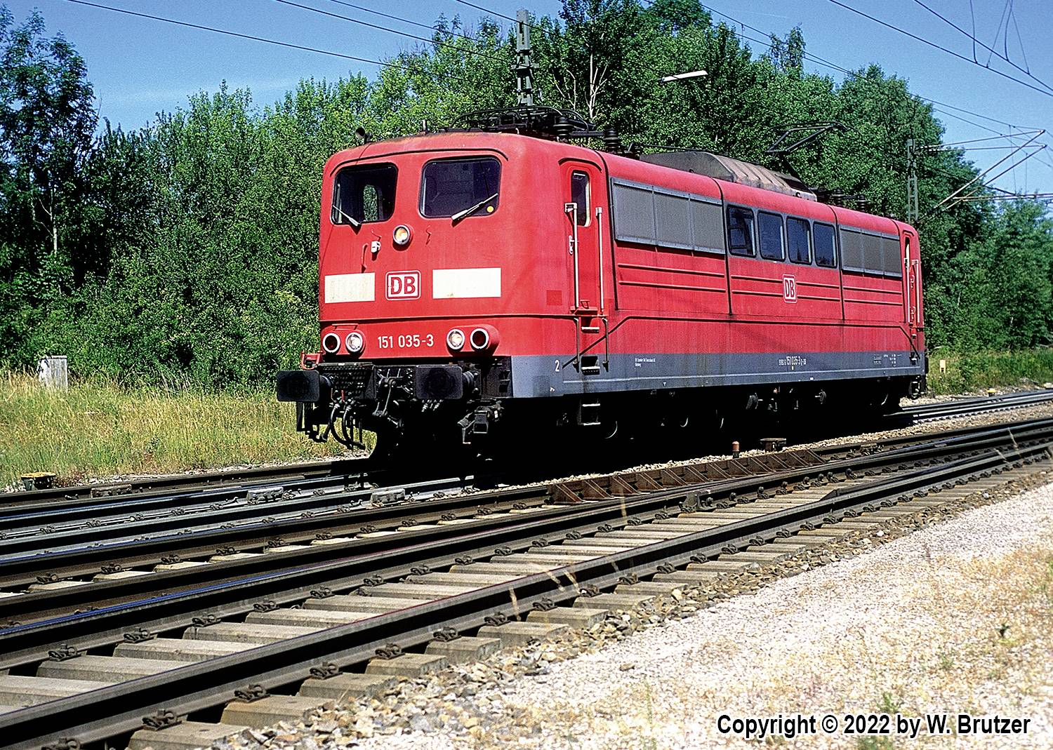 Roter Zug der Deutschen Bahn auf einem Gleis in einer ländlichen Umgebung bei sonnigem Wetter. Bäume im Hintergrund.