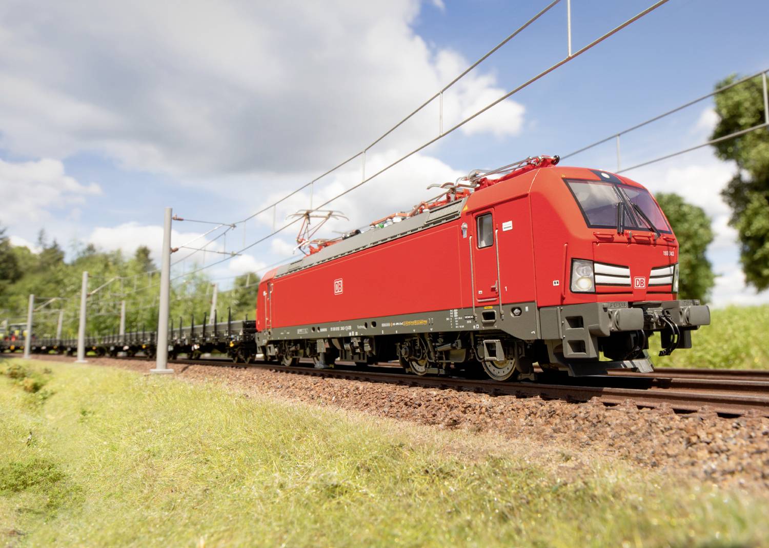 Ein roter Güterzug fährt auf einer zweigleisigen Bahnstrecke durch eine grüne Landschaft bei sonnigem Wetter.