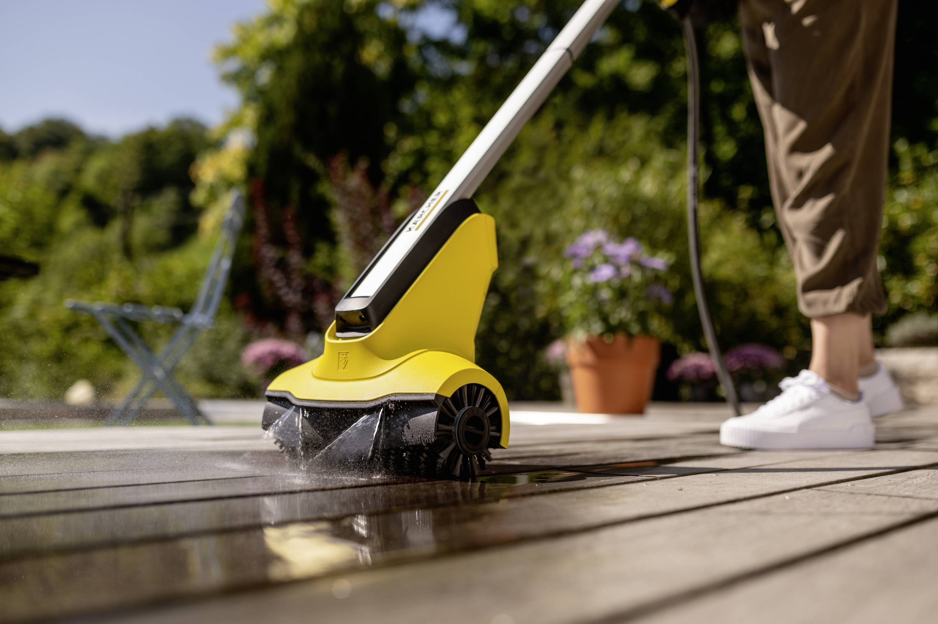 Person reinigt eine Holzterrasse mit einem gelben Hochdruckreiniger an einem sonnigen Tag, im Hintergrund Pflanzen und Möbel.