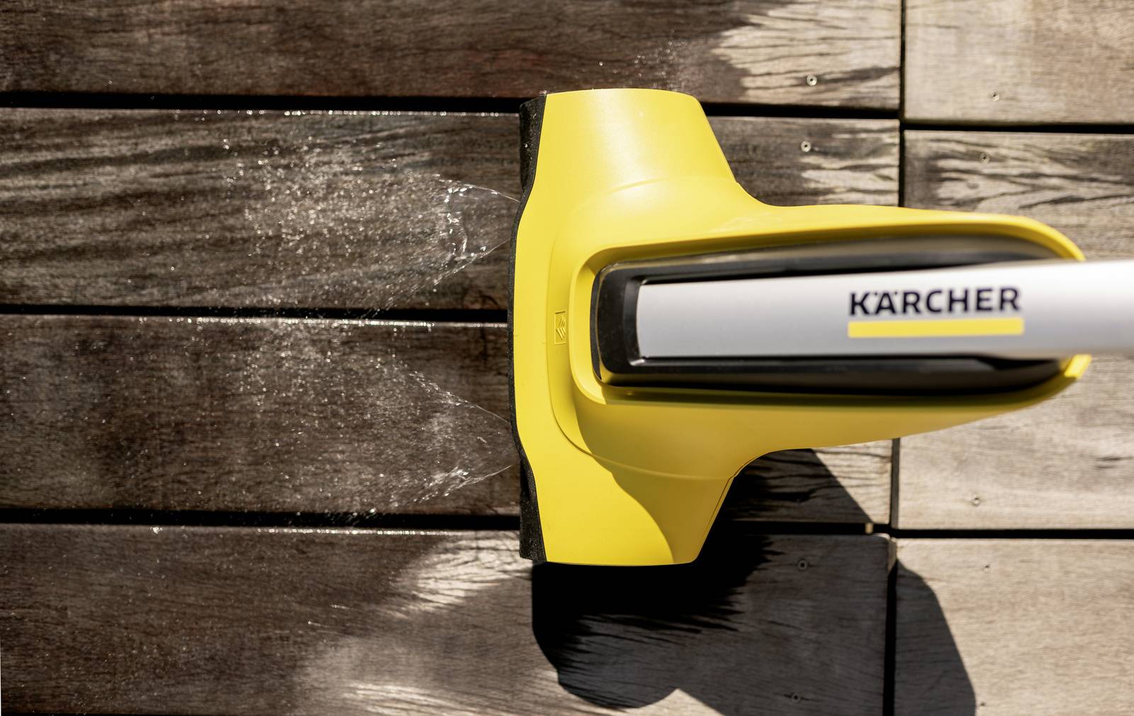 Ein gelber Hochdruckreiniger von Kärcher reinigt eine Holzterrasse. Links ist das Holz sauber, rechts noch schmutzig.