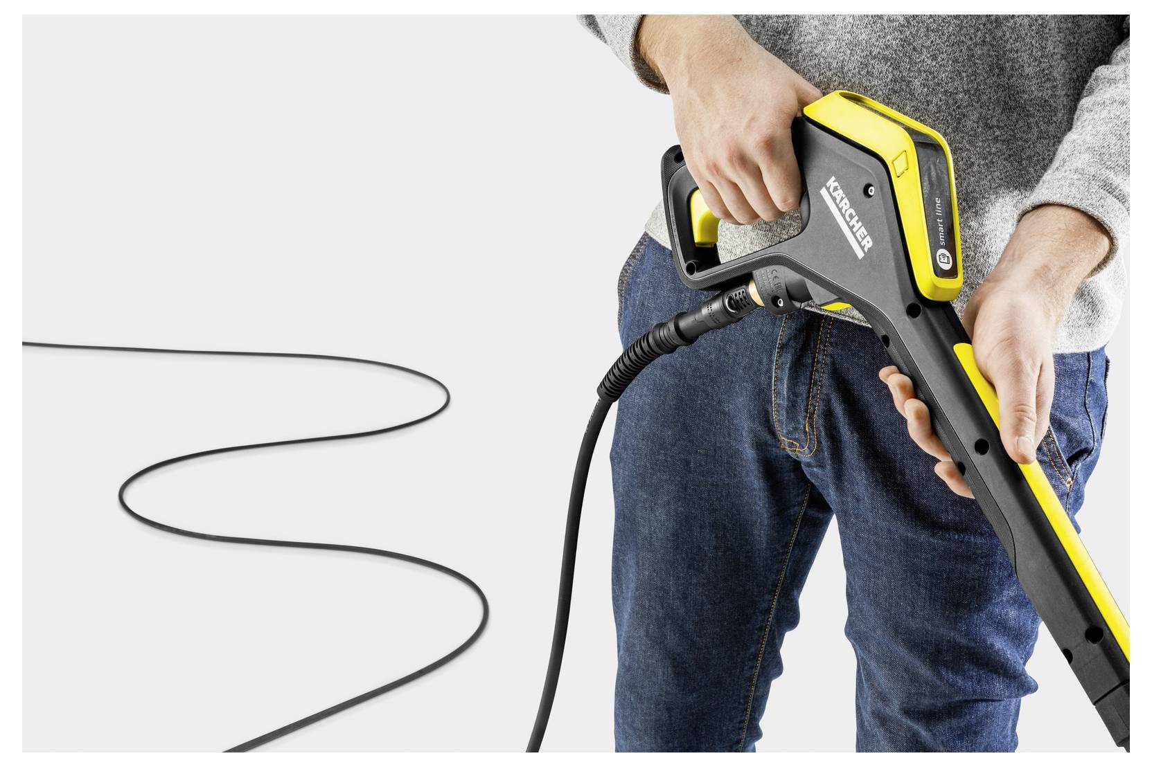 Eine Person, die einen Kärcher-Hochdruckreiniger-Griff hält, mit einem langen schwarzen Kabel, das sich über den Boden erstreckt, vor einem einfarbigen Hintergrund.