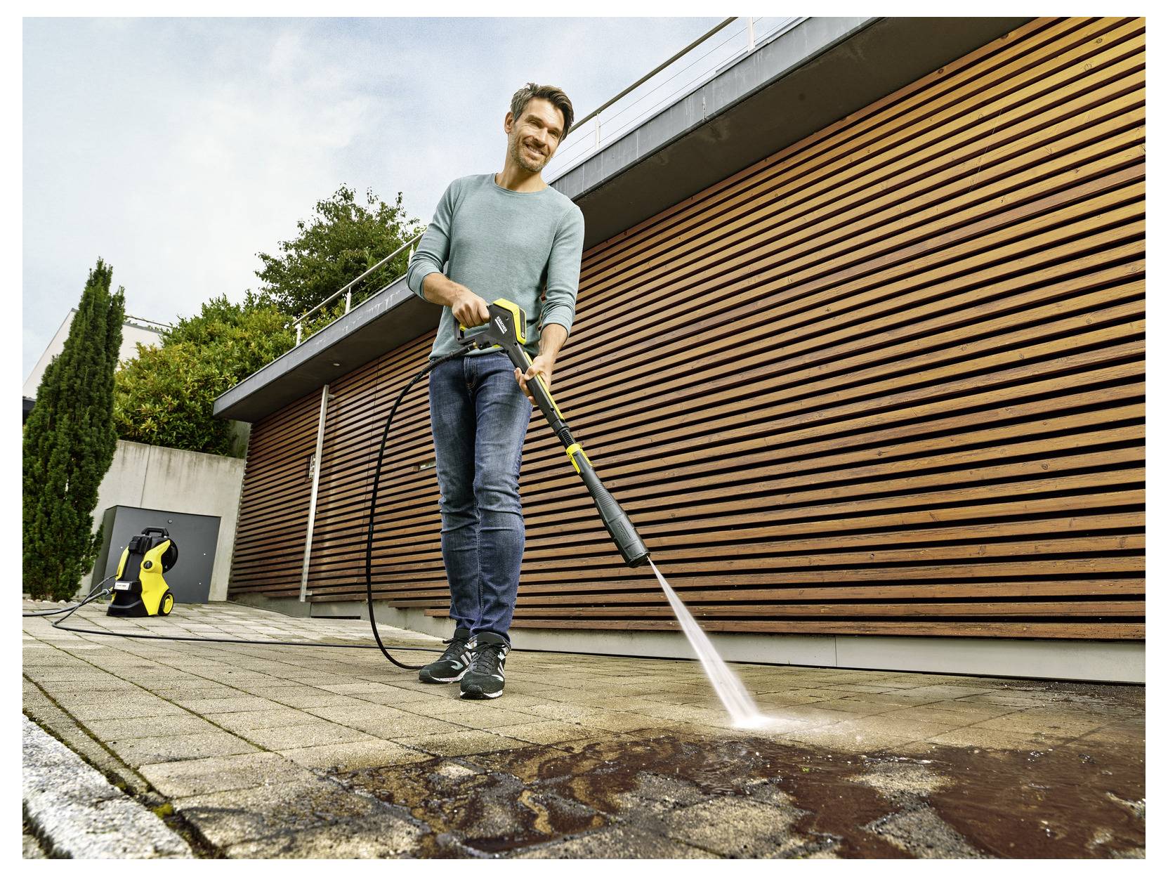 Eine Person reinigt eine Terrasse im Freien mit einem Hochdruckreiniger und entfernt dabei Schmutz. Sie benutzt einen gelben Hochdruckreiniger, der an einen Wasserschlauch angeschlossen ist.