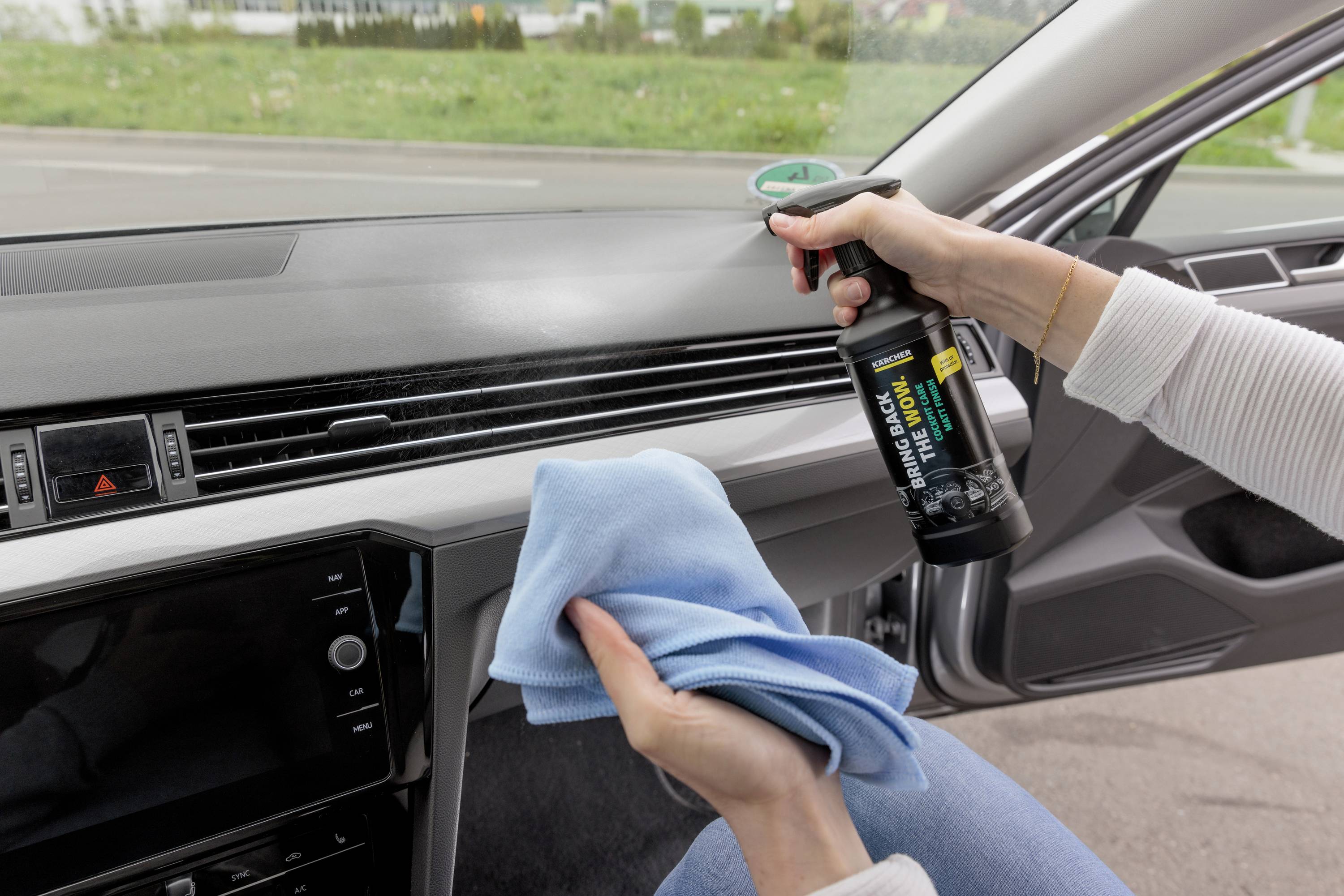 Person reinigt Autoinnenraum mit Sprühflasche und Tuch. Fokus liegt auf Armaturenbrettpflege.