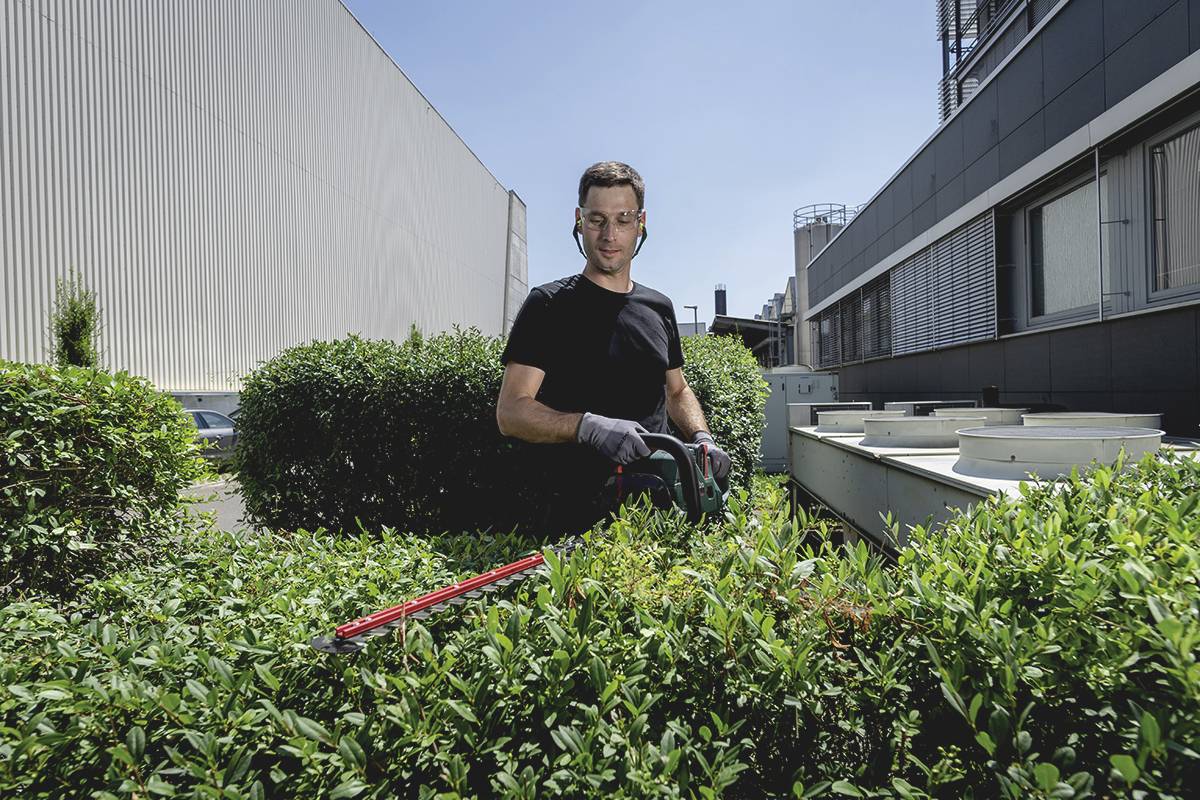 Ein Mann in einem schwarzen T-Shirt schneidet mit einer Heckenschere eine Hecke vor einem modernen Gebäude.