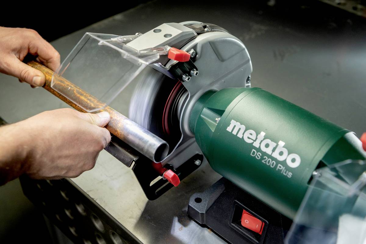 Person verwendet einen Metabo DS 200 Plus Schleifmaschine, um Rost von einem Metallrohr zu entfernen. Hände halten das Rohr stabil.