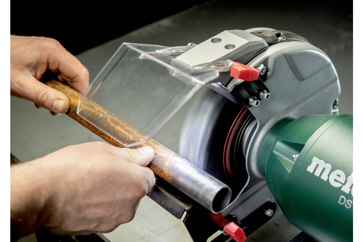 Person hält ein rostiges Rohr an ein Schleifgerät. Funken fliegen nicht, Schutzvorrichtung vorhanden. Metabo-Logo sichtbar.