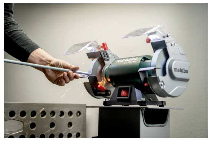 Eine Person verwendet einen Metabo Schleiftisch, um einen Stab zu schärfen, wobei Funken fliegen. Die Maschine ist grün mit Schutzvorrichtungen über den Schleifscheiben.