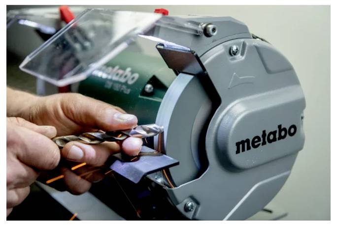 Eine Person benutzt einen Bohrerschärfer und hält einen Bohrer gegen das Schleifrad des Werkzeugs, um dessen Spitze zu verfeinern.