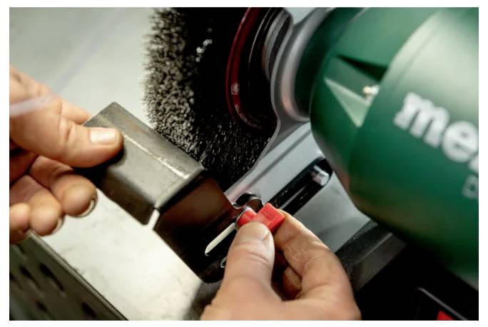 Hände, die eine Maschine mit einer Drahtbürste verwenden, um ein Metallobjekt zu polieren oder zu reinigen. Das Werkzeug hat ein grünes Gehäuse und die Maschine läuft.