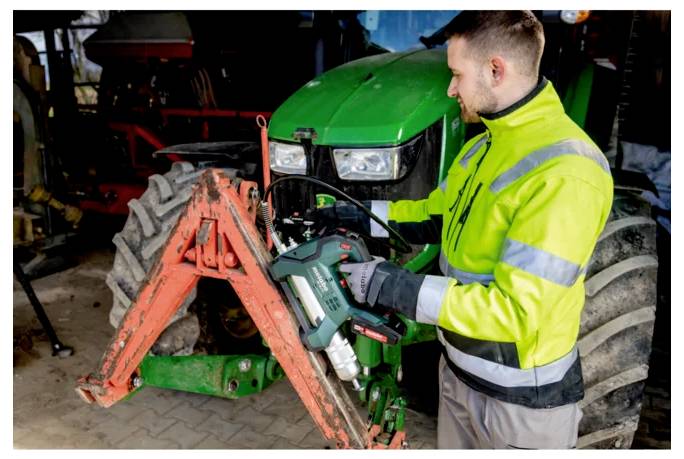 Ein Mechaniker in einer leuchtend farbigen Weste nutzt ein Elektrowerkzeug, um die Frontvorrichtung eines grünen Traktors in einer Werkstatt zu warten.