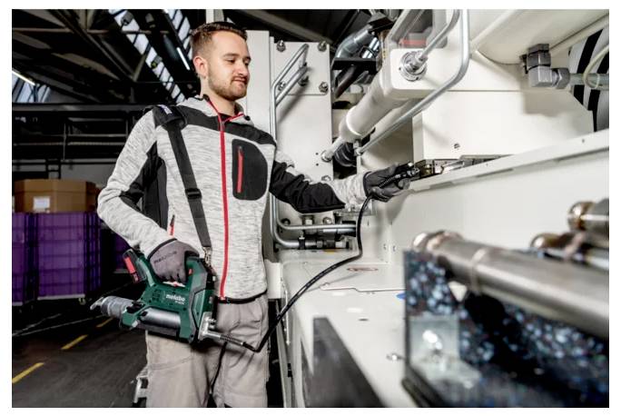 Ein Arbeiter in Schutzkleidung überprüft und bedient Maschinen in einer Industrieanlage und hält einen Elektroschrauber neben großen Rohren und Ausrüstungen.
