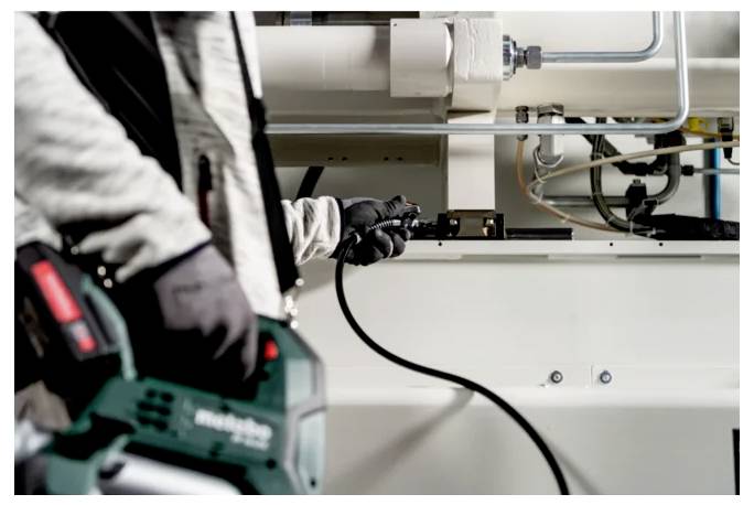 Arbeiter, der ein Elektrowerkzeug verwendet, um Maschinen in einer Industriewerkstatt einzustellen, mit sichtbaren Kabeln und Metallausrüstung.