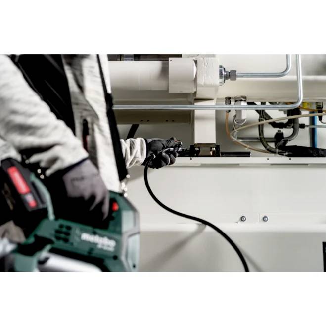 Eine Person in Arbeitshandschuhen hält ein Elektrowerkzeug und stellt eine Maschine in einer industriellen Umgebung mit sichtbaren Rohren und Metalloberflächen ein.