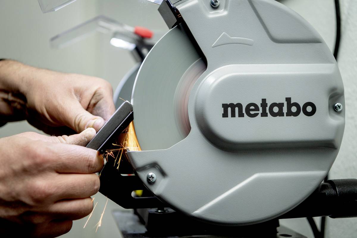 Eine Person schleift ein Metallwerkzeug mit einem Metabo-Schleifgerät, Funken fliegen. Fokus liegt auf der Schleifscheibe und den Händen.