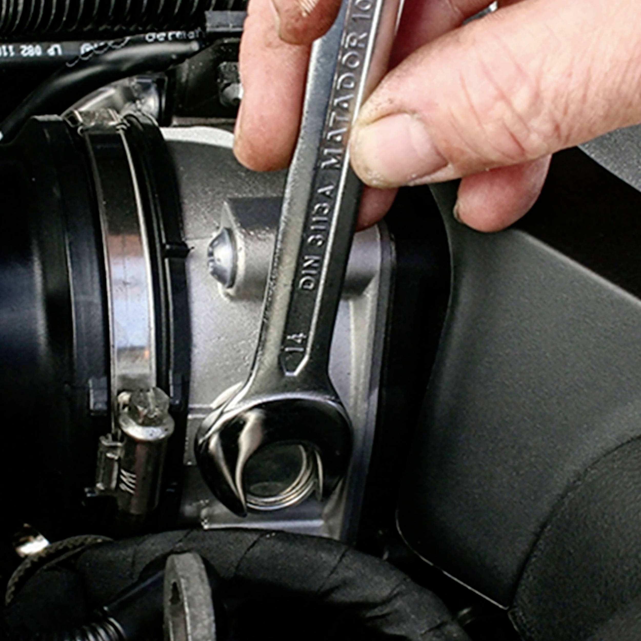 Eine Hand hält einen Schraubenschlüssel, der an einem Motorteil in einem Auto angebracht wird. Der Fokus liegt auf der Wartung.
