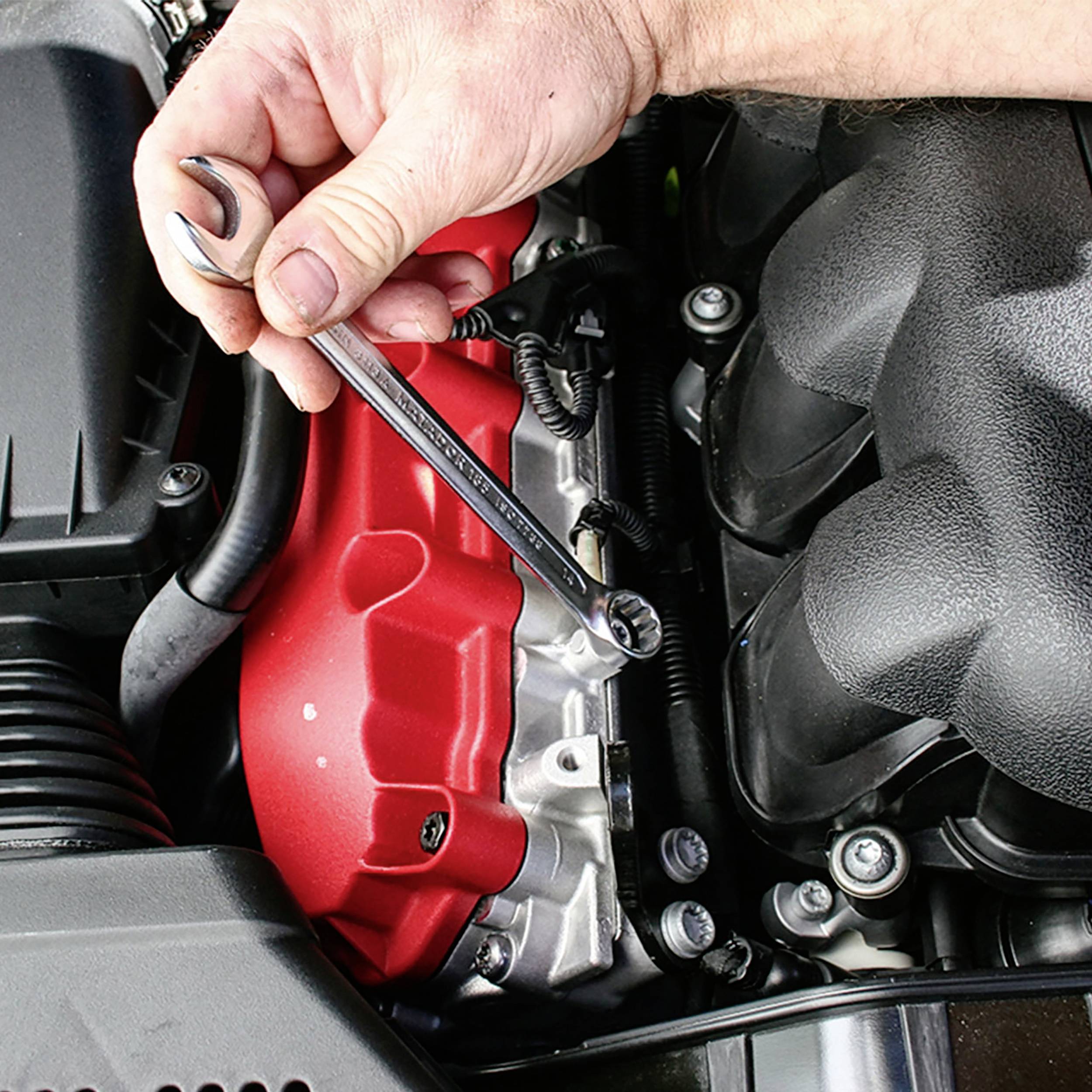 Eine Hand hält einen Schraubenschlüssel, der an einer Schraube eines Automotors arbeitet. Der Motor hat eine rote Abdeckung.