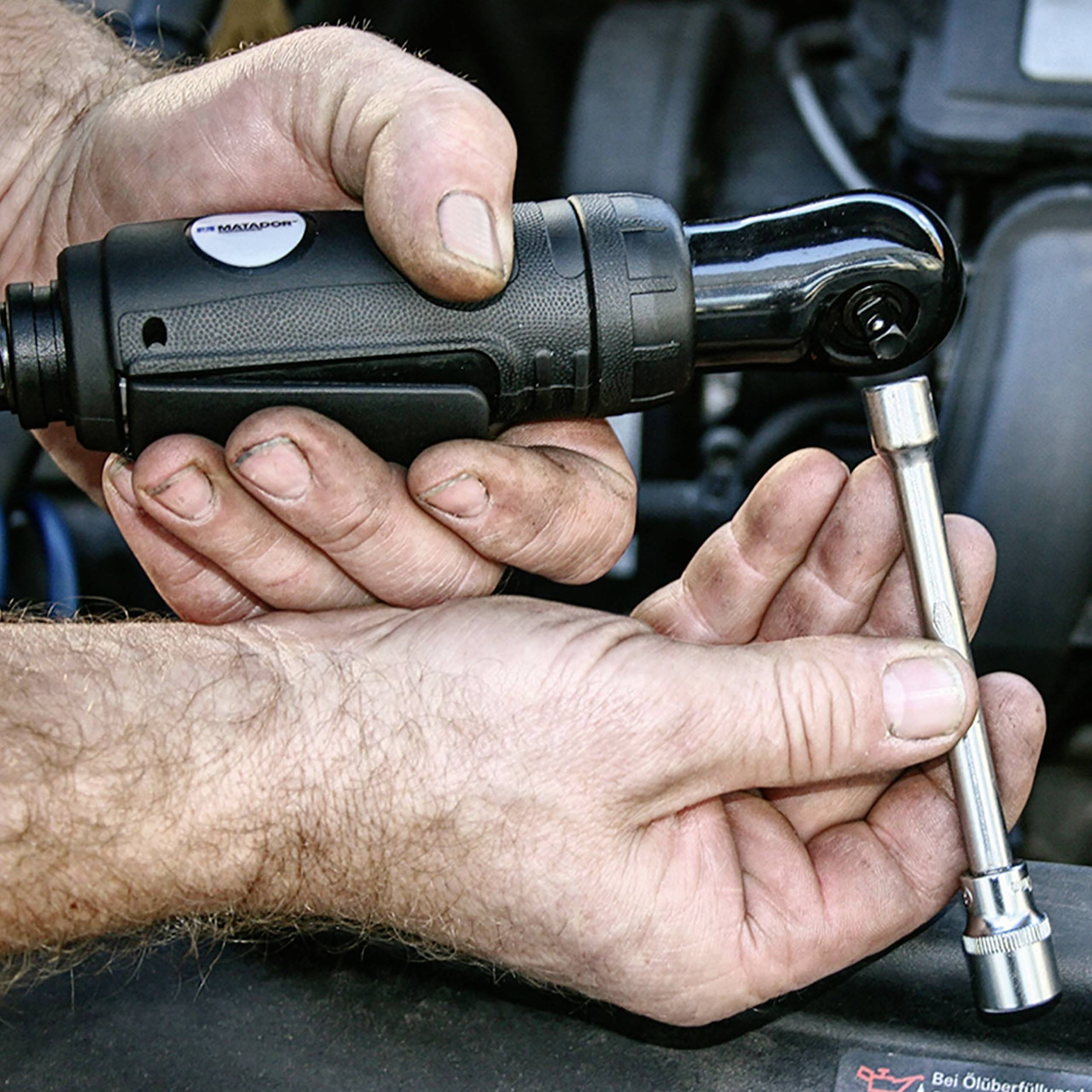 Mann hält einen elektrischen Schraubenschlüssel in der Hand, der eine Schraube anzieht oder löst. Hintergrund zeigt Teile eines Motors.