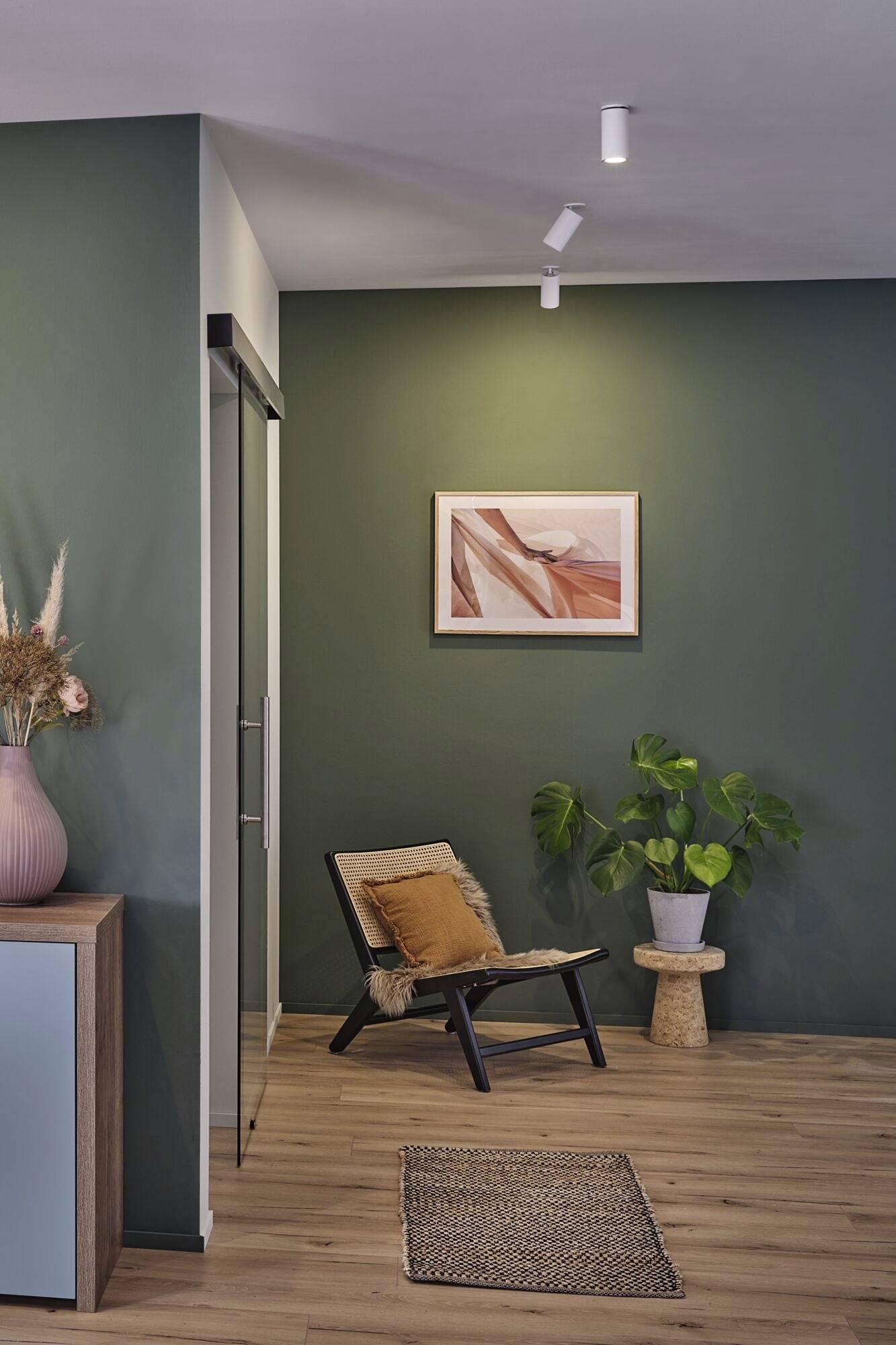 Ein Wohnzimmer mit grün gestrichener Wand, dekoriert mit einem gerahmten Bild. Ein Stuhl mit Kissen steht neben einer Pflanze.